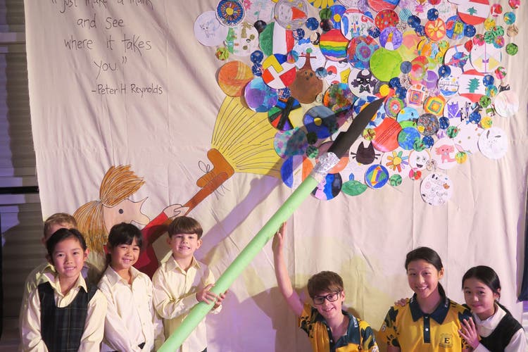 Photograph of students holding a giant paintbrush in front of a whole-school created artwork for an art show.